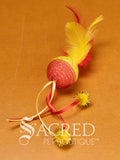 A single red and yellow mesh ball cat toy with ribbons and feathers on a wooden background with a white Sacred Pet Boutique watermark. 