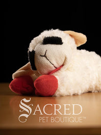 Two plush Lamb Chop sheep dog toys with red feet and tongues on a wooden surface, branded 'Sacred Pet Boutique'.