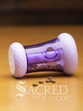 A purple tapered rolling cat toy with a treat dispensing hole in the side. The toy has cat kibble inside and some have escaped onto the wooden surface. A white Sacred Pet Boutique watermark.