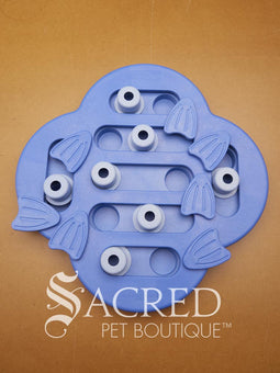 A blue Nina Ottosson Dog Hide N' Slide treat dispensing dog game with various compartments and flippers. The toy has a unique clover or cloud-like shape with channels and sliding pieces. It features multiple circular pale blue sliding blocks with small scent holes on top alongside several rotating flippers shaped like fish tails to cover hidden treat compartments. This is on a wooden background with a white Sacred Pet Boutique watermark. 