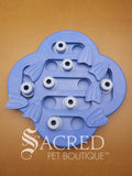A blue Nina Ottosson Dog Hide N' Slide treat dispensing dog game with various compartments and flippers. The toy has a unique clover or cloud-like shape with channels and sliding pieces. It features multiple circular pale blue sliding blocks with small scent holes on top alongside several rotating flippers shaped like fish tails to cover hidden treat compartments. This is on a wooden background with a white Sacred Pet Boutique watermark. 