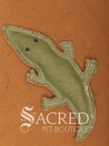 Corey the Crocodile is a green jute dog toy with multiple rows of stitching, on a wooden background. A white Sacred Pet Boutique watermark.