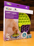 The Lickin' Layers food dispenser is shown in it's packaging, sitting on a wooden table with a wooden plank wall background. You can see two of the three layers of the toy in hexagonal and round designs.
