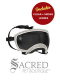 A pair of white protective goggles designed for dogs featuring a clear lens. The frame is fitted with a soft black foam padding along the inner edge for comfort and it has adjustable black nylon straps with a plastic buckle. The name Rex Specs is embossed on the upper right side of the frame. The goggles are on a white surface with a white background and above it is a circular red badge saying 'Includes clear + smoke lenses'. There is a grey Sacred Pet Boutique watermark at the bottom of the image. 
