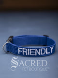 A blue dog collar with embroidered safety message "Friendly" in white. With a steel D-ring and black plastic side release clip. On a grey background with a Sacred Peet Boutique watermark.