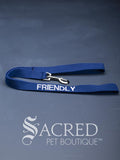 A blue dog leash with embroidered safety message "Friendly" in white. With a steel snap hook. On a grey background with a Sacred Peet Boutique watermark.