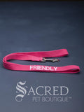 A Bright Pink dog leash with embroidered safety message "Friendly" in white. With a steel snap hook. On a grey background with a Sacred Peet Boutique watermark.