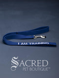 A blue dog leash with embroidered safety message "I am training" in white. With a steel snap hook. On a grey background with a Sacred Peet Boutique watermark.