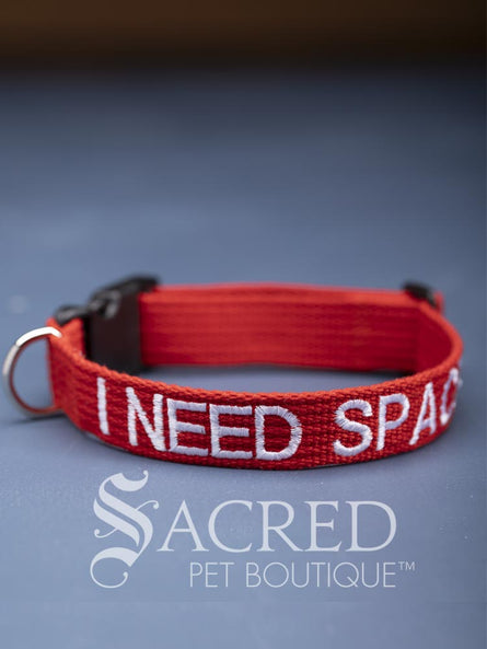 A bright red dog collar with embroidered safety message "I Need Space" in white. With a steel D-ring and black plastic side release clip. On a grey background with a Sacred Peet Boutique watermark.