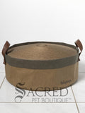 A round cardboard cat scratcher in a stylish, neutral beige fabric base cover with two dark brown leather look handles and the name ibiyaya printed on the side. It is sitting on a white tilled floor with a white background and a grey Sacred Pet Boutique watermark. 