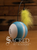A blue and white tumbling ball with a thin, flexible black wire that extends upwards from the top centre and has a green feather attached at the end of it. The brand Scream is printed in black on the white side of the ball. The toy is sitting on a wooden background with a white 'Sacred Pet Boutique' watermark. 