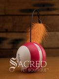A pink and white tumbling ball with a thin, flexible black wire that extends upwards from the top centre and has an orange feather attached at the end of it. The brand Scream is printed in black on the white side of the ball. The toy is sitting on a wooden background with a white 'Sacred Pet Boutique' watermark. 