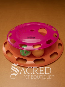 A close up shot of a cat toy consisting of a blue and yellow plastic tower with two balls and a spin top. One ball is an orange ping pong ball and the other is a green and white slotted lattice ball with an internal jingle bell. Sitting on a wooden background and a white Sacred Pet Boutique watermark. 
