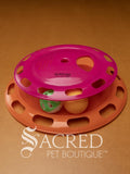 A cat toy consisting of a pink and orange plastic tower with two balls and a spin top. One ball is an orange ping pong ball and the other is a green and white slotted lattice ball with an internal jingle bell. Sitting on a wooden background and a white Sacred Pet Boutique watermark. 