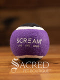 A single purple and black tennis ball sits on a wooden table with a wooden background. The ball is solid foam, so it won't pop, has dog friendly fuzz and does not squeak.