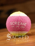 A single pink and green tennis ball sits on a wooden table with a wooden background. The ball is solid foam, so it won't pop, has dog friendly fuzz and does not squeak.