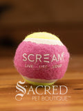 A single pink and green small tennis ball sits on a wooden table with a wooden background. The ball is solid foam, so it won't pop, has dog friendly fuzz and does not squeak.