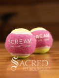 A set of two pink and green small tennis balls sit on a wooden table with a wooden background. The ball is solid foam, so it won't pop, has dog friendly fuzz and does not squeak.