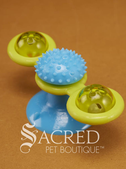 A vibrant blue and yellow rotatable windmill toy which includes two translucent side compartments in which one houses a bell and can both hold catnip or treats. At the base of the toy is a blue suction cup for attaching it to the floor or wall and in the centre there is a blue soft massager for self grooming. The toy is on a wooden background and has a white Sacred Pet Boutique watermark.