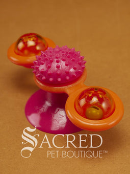 A spinning windmill cat toy with a blue spiky center and yellow edges, attached to a suction cup, displaying a blurred spinning effect to show how the toy spins. On a wooden background with a white Sacred Pet Boutique watermark. 