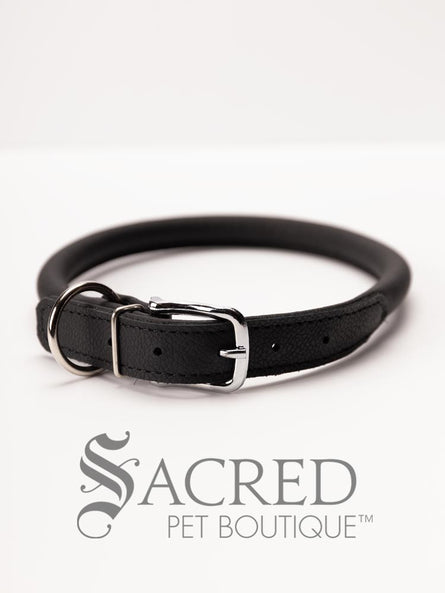 A black, round, soft leather dog collar with a metal buckle clasp. Our beautiful Soft Round (Rolled) leather collars are highly recommended for long haired dogs. Round or rolled collars reduce the amount of matting, tangling and flattening of the dogs fur. All collars are hand made and stitched with wax thread, from the finest quality soft leather wrapped and stitched around a nylon inner cord for added strength.Image on a white background, with the Sacred Pet Boutique logo.