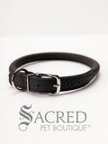 A black, round, soft leather dog collar with a metal buckle clasp. Our beautiful Soft Round (Rolled) leather collars are highly recommended for long haired dogs. Round or rolled collars reduce the amount of matting, tangling and flattening of the dogs fur. All collars are hand made and stitched with wax thread, from the finest quality soft leather wrapped and stitched around a nylon inner cord for added strength.Image on a white background, with the Sacred Pet Boutique logo.