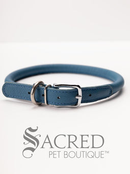 A black, round, soft leather dog collar with a metal buckle clasp. Our beautiful Soft Round (Rolled) leather collars are highly recommended for long haired dogs. Round or rolled collars reduce the amount of matting, tangling and flattening of the dogs fur. All collars are hand made and stitched with wax thread, from the finest quality soft leather wrapped and stitched around a nylon inner cord for added strength.Image on a white background, with the Sacred Pet Boutique logo.