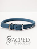 A blue, round, soft leather dog collar with a metal buckle clasp. Our beautiful Soft Round (Rolled) leather collars are highly recommended for long haired dogs. Round or rolled collars reduce the amount of matting, tangling and flattening of the dogs fur. All collars are hand made and stitched with wax thread, from the finest quality soft leather wrapped and stitched around a nylon inner cord for added strength.Image on a white background, with the Sacred Pet Boutique watermark.