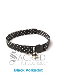 A cat collar is shown against a plain white background. The pattern of the fabric is pink with white polker dots. The collar has a black plastic breakaway buckle and adjuster, and a silver bell on a silver split ring.