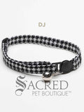 A Black and white houndstooth patterned soft fabric cat collar with black plastic hardware and a silver bell with a safety breakaway buckle on a white background. A Sacred Pet Boutique watermark.