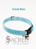 An aqua blue crack patterned soft fabric cat collar with black plastic hardware and a silver bell with a safety breakaway buckle on a white background. A Sacred Pet Boutique watermark.