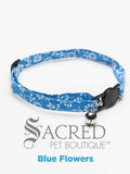 A blue with white flowers patterned soft fabric cat collar with black plastic hardware and a silver bell with a safety breakaway buckle on a white background. A Sacred Pet Boutique watermark.