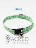 A Green with white daisy flowers soft fabric cat collar with black plastic hardware and a silver bell with a safety breakaway buckle on a white background. A Sacred Pet Boutique watermark.