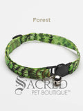 A green leaf patterned soft fabric cat collar with black plastic hardware and a silver bell with a safety breakaway buckle on a white background. A Sacred Pet Boutique watermark.