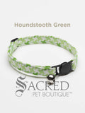 A green houndstooth patterned soft fabric cat collar with black plastic hardware and a silver bell with a safety breakaway buckle on a white background. A Sacred Pet Boutique watermark.