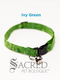 A green ivy patterned soft fabric cat collar with black plastic hardware and a silver bell with a safety breakaway buckle on a white background. A Sacred Pet Boutique watermark.