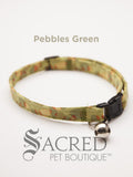 A green pebble patterned soft fabric cat collar with black plastic hardware and a silver bell with a safety breakaway buckle on a white background. A Sacred Pet Boutique watermark.