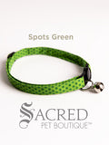 A green spot patterned soft fabric cat collar with black plastic hardware and a silver bell with a safety breakaway buckle on a white background. A Sacred Pet Boutique watermark.