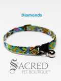 A multicoloured diamond pattern soft fabric cat collar with black plastic hardware and a silver bell with a safety breakaway buckle on a white background. A Sacred Pet Boutique watermark.