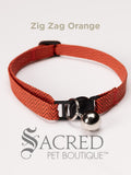 An orange and black zigzag patterned soft fabric cat collar with black plastic hardware and a silver bell with a safety breakaway buckle on a white background. A Sacred Pet Boutique watermark
