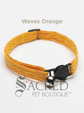An orange wavy lines soft fabric cat collar with black plastic hardware and a silver bell with a safety breakaway buckle on a white background. A Sacred Pet Boutique watermark