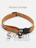 An orange white red green blue striped soft fabric cat collar with black plastic hardware and a silver bell with a safety breakaway buckle on a white background. A Sacred Pet Boutique watermark