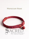 A pink red orange and aqua blue Moroccan patterned soft fabric cat collar with black plastic hardware and a silver bell with a safety breakaway buckle on a white background. A Sacred Pet Boutique watermark.