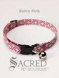 A pink retro geometric patterned soft fabric cat collar with black plastic hardware and a silver bell with a safety breakaway buckle on a white background. A Sacred Pet Boutique watermark.