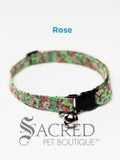 A pink rose pattern on a green background soft fabric cat collar with black plastic hardware and a silver bell with a safety breakaway buckle on a white background. A Sacred Pet Boutique watermark.
