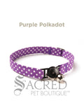 A purple Polka Dot soft fabric cat collar with black plastic hardware and a silver bell with a safety breakaway buckle on a white background. A Sacred Pet Boutique watermark.