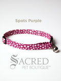 A purple with white spot patterned soft fabric cat collar with black plastic hardware and a silver bell with a safety breakaway buckle on a white background. A Sacred Pet Boutique watermark. 