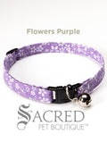 A purple with white flowers patterned soft fabric cat collar with black plastic hardware and a silver bell with a safety breakaway buckle on a white background. A Sacred Pet Boutique watermark.