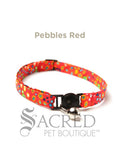 A red pebble patterned soft fabric cat collar with black plastic hardware and a silver bell with a safety breakaway buckle on a white background. A Sacred Pet Boutique watermark.
