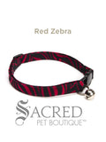 A red zebra patterned soft fabric cat collar with black plastic hardware and a silver bell with a safety breakaway buckle on a white background. A Sacred Pet Boutique watermark.